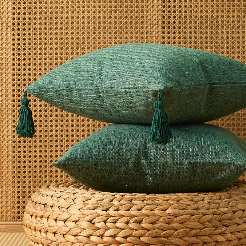 A deep green linen throw pillow with tasseled trim rests on a woven natural fiber surface against a textured wall.