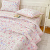 A pink and white floral quilted bedspread is draped over a bed with pink accents and pillows.