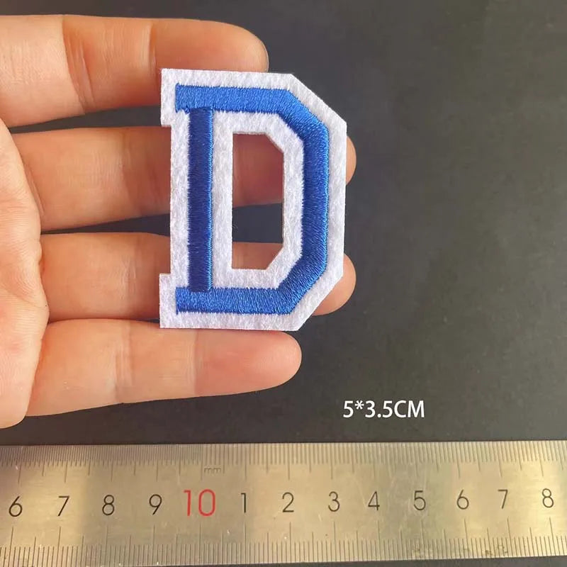 A hand holds a blue alphabet embroidery iron-on patch shaped like the letter D, measured against a ruler.