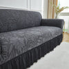 A charcoal gray jacquard stretch sofa cover featuring a floral pattern and a ruffled hem.