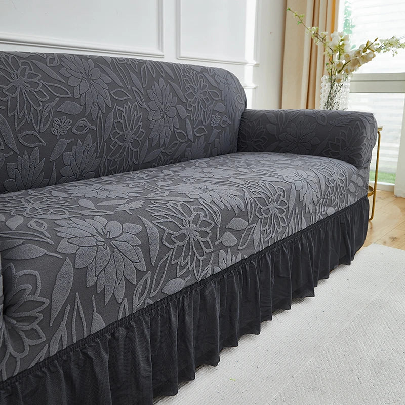 A charcoal gray jacquard stretch sofa cover featuring a floral pattern and a ruffled hem.