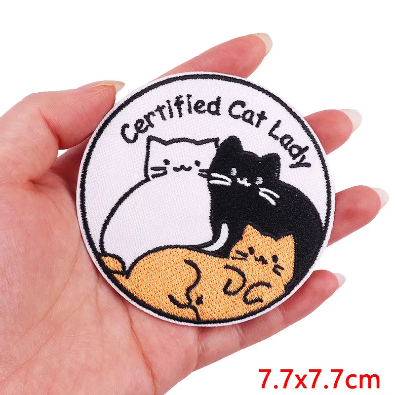 A hand holds a circular embroidered patch featuring cartoon cats, including a black cat and a white cat.