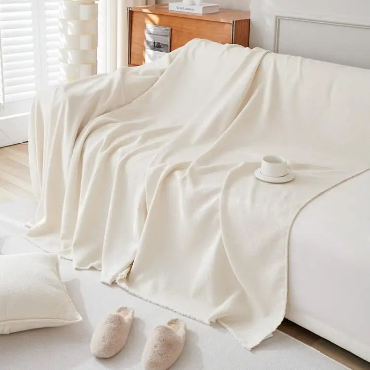 A cream anti-scratch sofa cover draped over a white sofa with a small cup and slippers nearby.