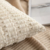 A cream-colored knitted throw pillow cover with a textured, repeating stripe pattern resting on a beige woven fabric.
