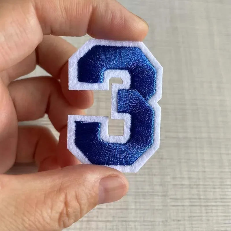 A hand holds a navy blue embroidered number 3 patch with a white border.