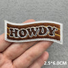 A brown embroidered patch spelling HOWDY is held in a hand against a gray background.