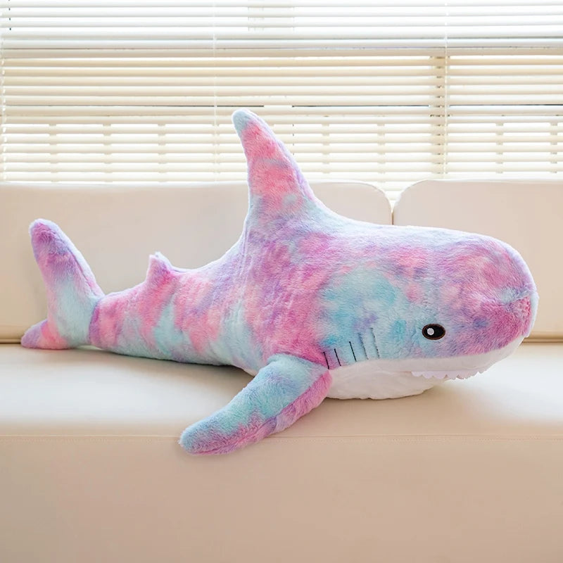 A pink and blue giant shark plush toy lying on a light-colored sofa.