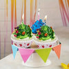 A tiered stand holds two cupcakes with green frosting and lit candles, set against a colorful striped background.
