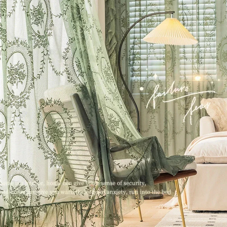 A sheer floral lace curtain panel in sage green hangs next to a wooden chair and a light-colored bed.