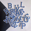 A collection of blue and white alphabet embroidery iron-on patches scattered on a dark surface.