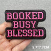 Black embroidered patch with pink text reading BOOKED BUSY BLESSED being held in a hand