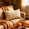 A cozy living room scene featuring a beige sofa with an orange knitted throw and a cream fuzzy maple leaf throw pillow.
