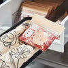 A clear plastic bag of pinkish-red floral heat-resistant oven mitts is placed on a quilted fabric liner in a baking tool hold