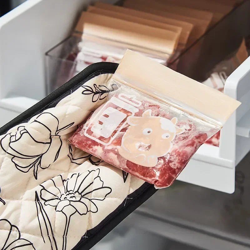 A clear plastic bag of pinkish-red floral heat-resistant oven mitts is placed on a quilted fabric liner in a baking tool hold