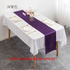 A white tablecloth with a purple satin table runner, gold cutlery, and pink floral arrangement on a dining table.