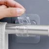 A hand is using a self-adhesive curtain rod clip to attach a clear plastic clip onto a silver curtain rod.