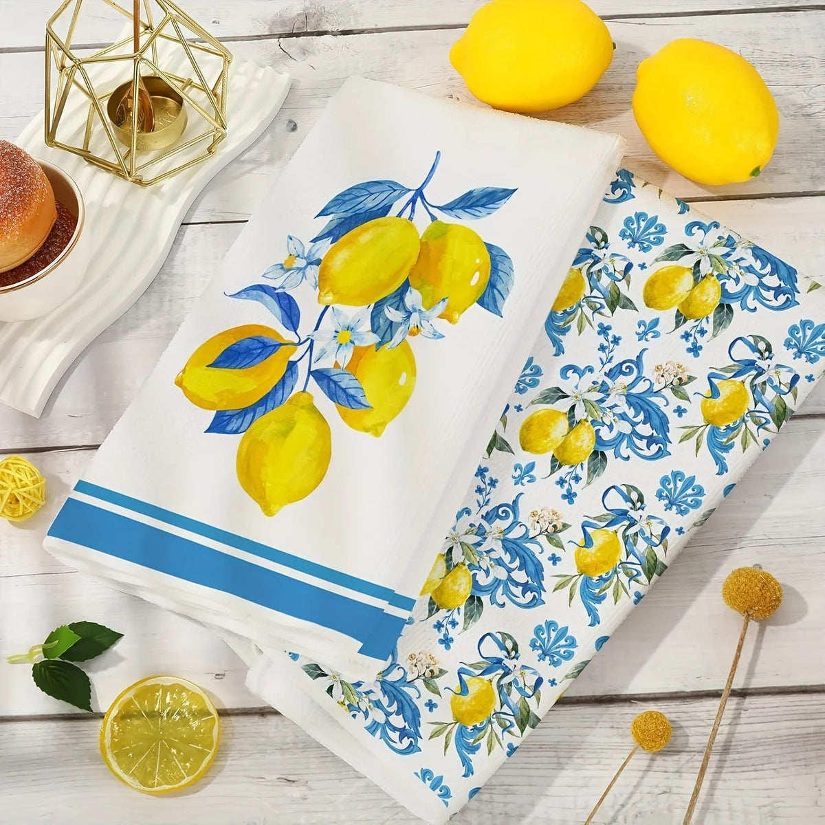 Two blue-edged lemon kitchen towels are displayed on a white wooden surface with fresh lemons.