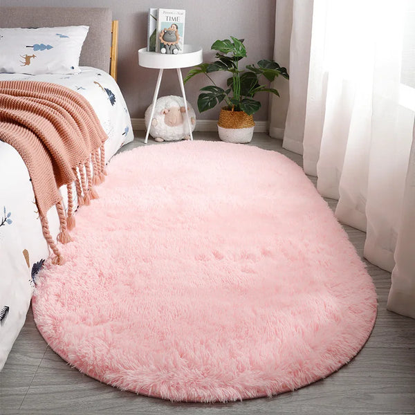 A round, fluffy pink plush floor mat placed on a wooden floor next to a bed and a potted plant.