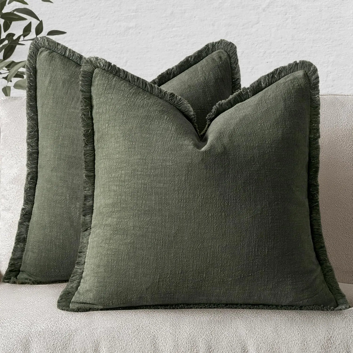 A dark olive green linen throw pillow with fringe resting on a light gray sofa.