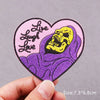 A pink heart-shaped embroidered patch featuring a yellow skull wearing a purple hood and the text Live Laugh Love