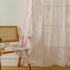 A sheer pink floral lace curtain hangs in a window, draped over a wooden chair and table.