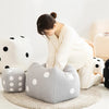 A woman is sitting on a beige floor cushion next to several gray and white plush dice cube cushions.