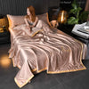 A luxurious satin pink ice silk blanket set is draped on a bed, next to a dark bedside table and a gold lamp.