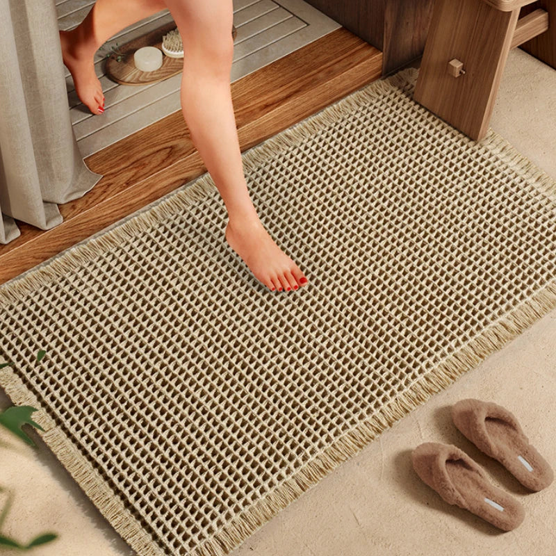 A person's bare foot steps onto a beige waffle-patterned non-slip bath mat on a wooden floor.