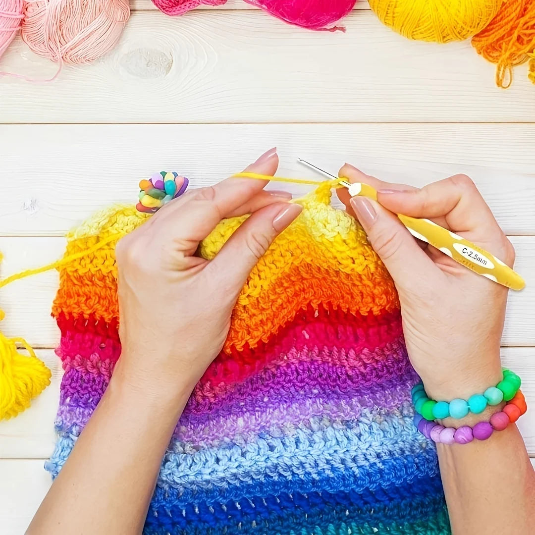 Hands are using a crochet hook to work on a brightly colored, rainbow-striped knitted or crocheted fabric.